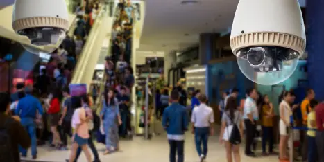 A security camera in a crowded shopping mall, symbolizing surveillance technology and the risks of facial recognition errors leading to wrongful arrests.