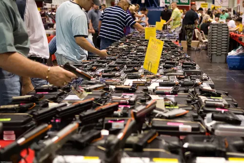 Gun show with tables full of firearms and multiple potential customers inspecting the weapons